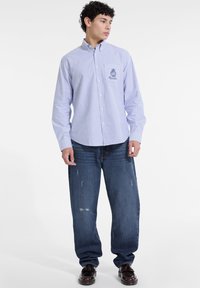 Camisa de botones con rayas azules y blancas, mangas largas, cuello puntiagudo y un logo bordado. Lleva con unos jeans de mezclilla holgados.