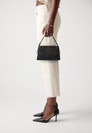 Sac à main en cuir noir avec une finition texturée, ornée d'un motif géométrique doré. Le modèle porte un jean crème et des escarpins noirs en vernis.