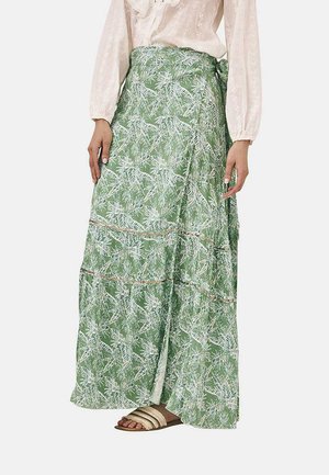 Larga falda maxi en capas de color verde con un patrón de hojas blancas, con un diseño envolvente y sutiles acentos dorados. Combinada con sandalias de color claro.