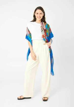 Lächelnde Frau steht, trägt weißes T-Shirt, weiße Hose mit hoher Taille, schwarze Sandalen und einen bunten blauen Blumenschal über den Schultern drapiert.