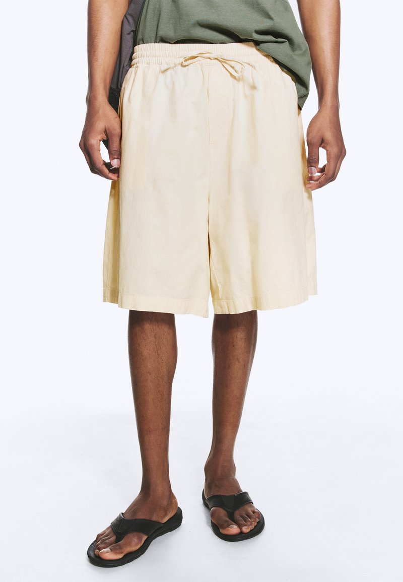Personne portant un short beige à cordon, un t-shirt vert et des tongs noires, debout devant un fond uni.