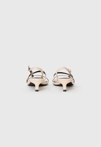 Et par beige stroppsandaler med lave kittenhæle, set fra bagsiden på en ensfarvet hvid baggrund.