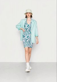 Blazer oversized verde menta sobre um vestido justo com padrão de espiral multi-colorido. O modelo usa um chapéu tipo balde claro e botas de cano curto brancas. Texturas suaves visíveis.