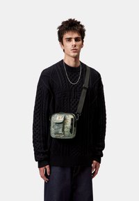 Bolso cruzado con un patrón de camuflaje, tapa frontal, correas de tela y un bolsillo con cremallera. Lleva puesto sobre un suéter de punto negro texturizado.