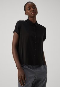 Kortermet svart skjorte med knapper og krage, glattes tekstur og subtil glans, kombinert med grå bukser. Modellen poserer rett.