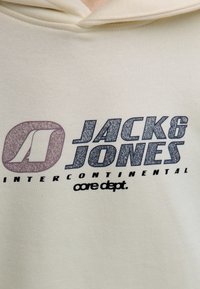 Un gros plan sur un hoodie beige affichant le logo "JACK & JONES" et le texte "INTERCONTINENTAL" en bleu foncé et marron.