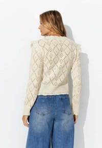 Pull en crème tricoté avec des motifs en losange, des bords festonnés et des épaules volantes, associé à un jean bleu taille haute.
