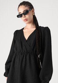 Melna, garām piedurknēm tērps ar V veida dekoltē un strukturētu audumu. Elastīga jostasvieta, plūstoša silueta, kombinēts ar taisnstūra melnām saulesbrillēm.