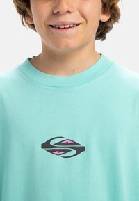 Camiseta de color azul aqua clara hecha de algodón, con un logo negro con detalles en rosa en la parte delantera. Diseño de cuello redondo y mangas cortas.