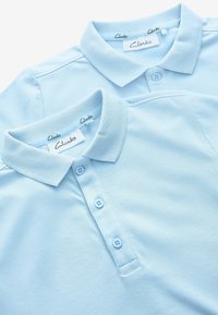 Dos camiseta polo azul claro hechas de tejido texturizado, con un cómodo cierre de tres botones y un cuello doblado. Marca Clarks visible en el interior.