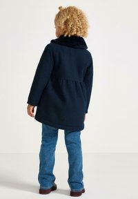 Bambino con capelli ricci che indossa un cappotto blu navy scuro con colletto di pelliccia, jeans blu e scarpe scure, in piedi con le spalle rivolte verso di noi su uno sfondo semplice.