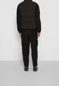 Schwarze Steppweste mit hohem Kragen, getragen über einem strukturierten schwarzen Cord-Outfit. Enthält Cargo-Taschen und schwarze klobige Stiefel.