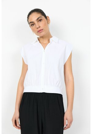 Woman with dark hair tied back wears a sleeveless white button-up shirt and loose black pants, standing against a plain white background.