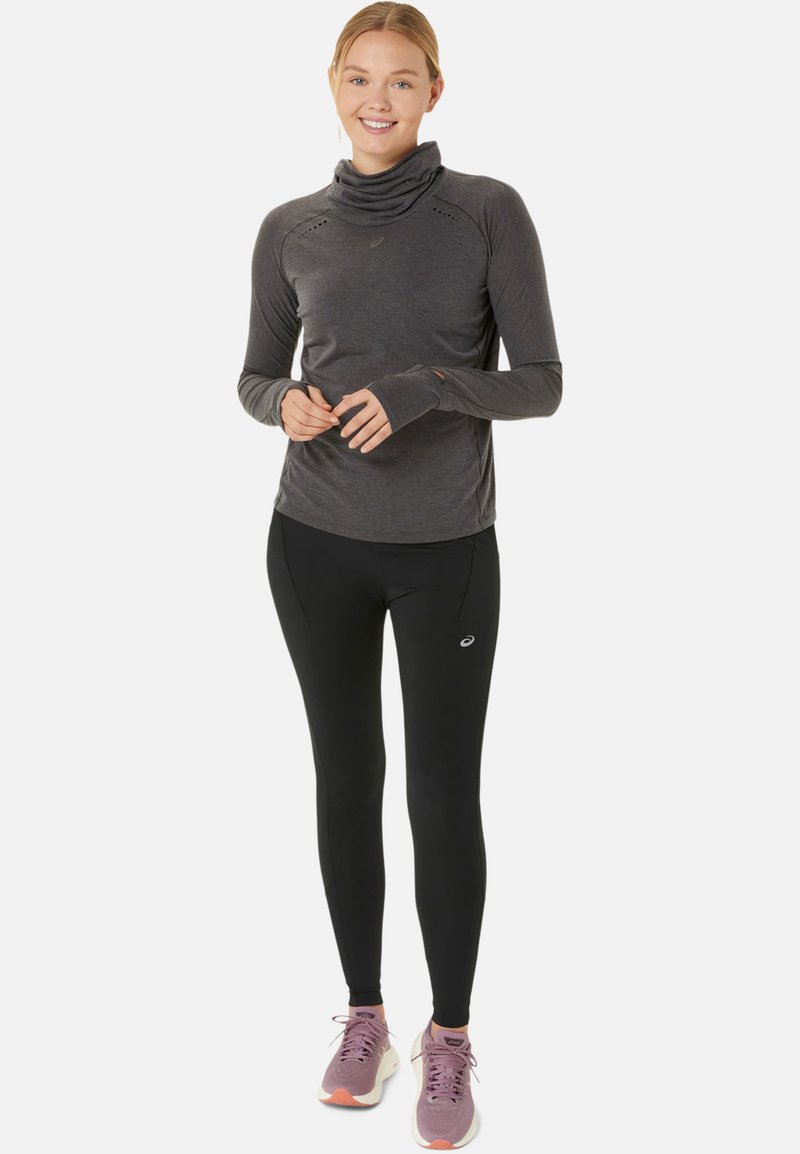 Haut de sport à manches longues, gris foncé avec un col cowl, associé à un legging noir et des baskets roses, arborant un logo sur le pantalon.