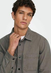 Camisa abotonada gris claro de tela suave con cuello de punto, un bolsillo en el pecho y botones negros, llevada sobre una camisa blanca.