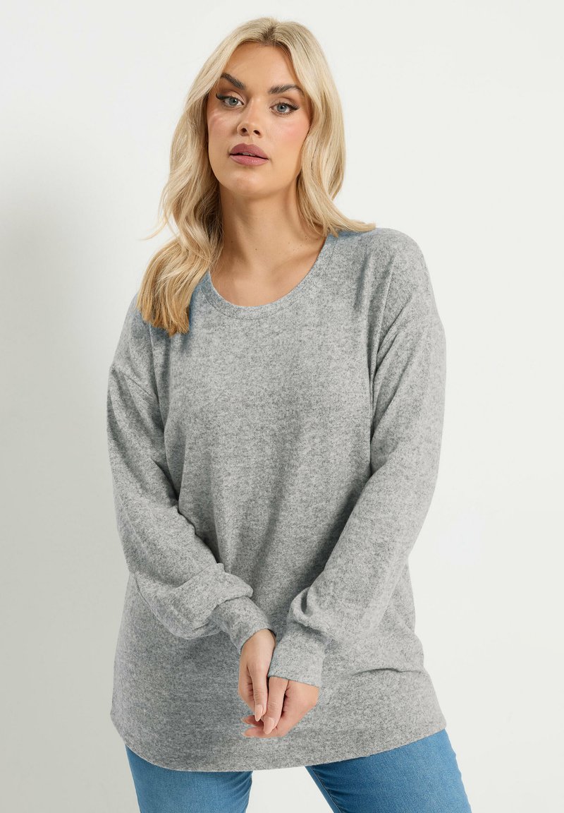 Donna bionda che indossa un maglione grigio a maniche lunghe e jeans blu, posa con le mani leggermente intrecciate davanti a uno sfondo semplice.