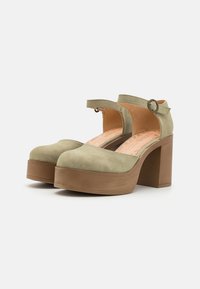 Tacchi a piattaforma verde in suede con punta rotonda, cinturino alla caviglia e un solido tacco a blocco marrone. Superficie texturizzata con una finitura liscia.