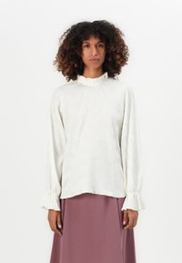 Donna con capelli ricci che indossa una blusa bianca a maniche lunghe con texture, con polsini arricciati e colletto alto, abbinata a una gonna color malva.