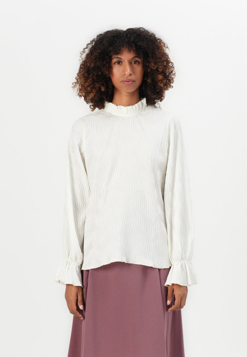 Donna con capelli ricci che indossa una blusa bianca a maniche lunghe con texture, con polsini arricciati e colletto alto, abbinata a una gonna color malva.