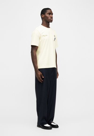 Άνδρας όρθιος με κρεμ μπλούζα με γραφικά, μαύρο-γκρι ριγέ φαρδιά παντελόνια και μαύρα και λευκά μοκασίνια, σε απλό λευκό φόντο.