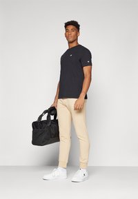 Black short-sleeve t-shirt with small logo, beige tapered joggers, white sneakers, and a black duffel bag with shoulder strap.