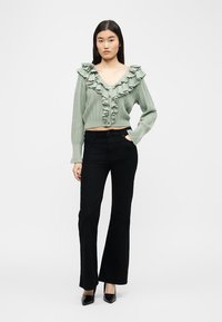 Γυναίκα με ανοιχτό πράσινο ζακέτα με κουμπιά και βολάν, και μαύρο ψηλόμεσο καμπάνα παντελόνι, στέκεται με το ένα χέρι στην τσέπη.