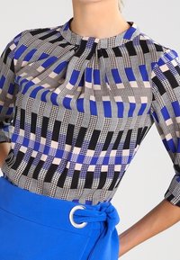 Kvinna som bär en mönstrad blus med svarta, blå och beige geometriska former, matchad med en ljusblå kjol som har ett silverringbälte.