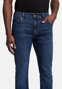 Ciemnoniebieskie dżinsy z prostymi nogawkami, charakteryzujące się klasycznym pięciopunktowym wzornictwem, żółtymi szwami, metalowym zapięciem na guziki oraz gładką fakturą.