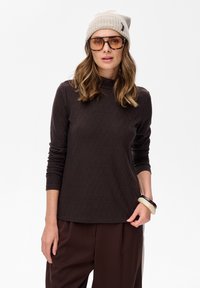 Frau trägt eine beige Strickmütze, übergroße Brille mit Schildpatt-Muster, dunkeltexturiertes Langarmoberteil, braune Hose und zwei Armbänder an einem Handgelenk.