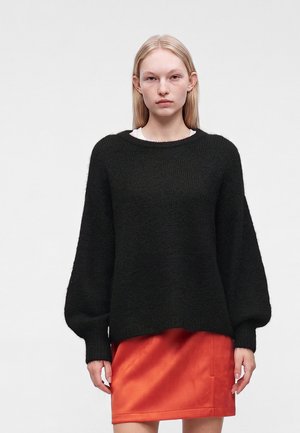 Jeune femme aux cheveux blonds et raides porte un pull en maille noir à manches ballons et une jupe courte orange vif sur un fond uni.
