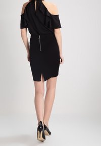 Vestido negro con mangas de volantes off-the-shoulder y forma ajustada. Presenta un cierre de cremallera en la parte posterior y una abertura en el dobladillo. Zapatos clásicos de tacón.