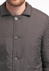 Prošívaná bunda tmavě šedé barvy s límcem, se čtyřmi černými knoflíky a viditelným prošitím. Hladká, lesklá textura.