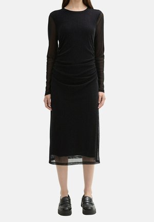 Vrouw draagt een zwarte midi-jurk met lange mouwen met doorschijnende mouwen en zoom, gecombineerd met zwarte chunky loafers, staand tegen een lichte achtergrond.