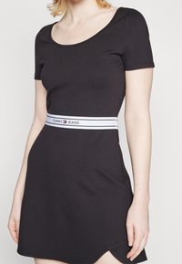 Nainen, jolla on päällään lyhythihainen musta mekko, jossa on valkoinen vyötärönauha, joka on merkitty "Tommy Jeans", ja helman lähellä on pieni halkio.