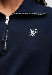 Hanorac bleumarin cu fermoar argintiu și inițialele albe "FH" brodate pe partea stângă a pieptului, purtat de o persoană cu păr creț închis.