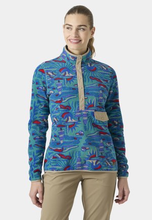 Mujer sonriente con un forro polar azul con estampado, con tapeta con botones de presión y bolsillo en color tostado, combinado con pantalones beige sobre un fondo liso.