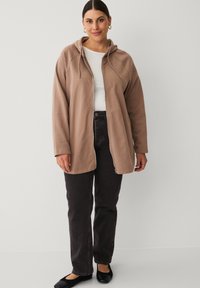 Light brown zip-up hoodie with a drawstring hood, relaxed fit, and long sleeves, paired with dark denim jeans and black flat shoes.