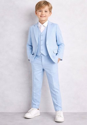 Jeune garçon en costume bleu clair avec gilet, chemise blanche et baskets blanches, debout, souriant sur un fond uni.