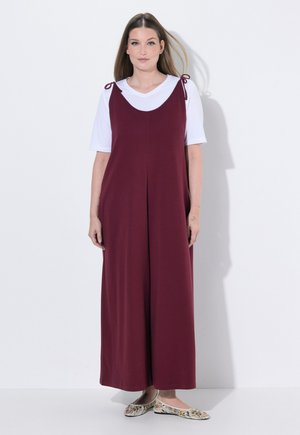 Kvinde, der står iført en hvid kortærmet skjorte under en ærmeløs, mørkerød jumpsuit med bundne skulderstropper og blomstrede flade sko.