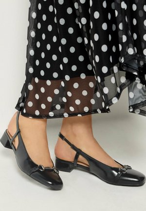 Füße tragen schwarze Slingback-Schuhe mit niedrigem Absatz unter einem durchsichtigen schwarzen Rock mit großen weißen Punkten.