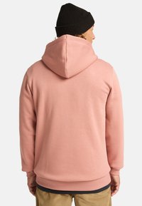 Pink hooded sweatshirt with a textured fabric, relaxed fit, and ribbed cuffs. Worn with a black knitted beanie.