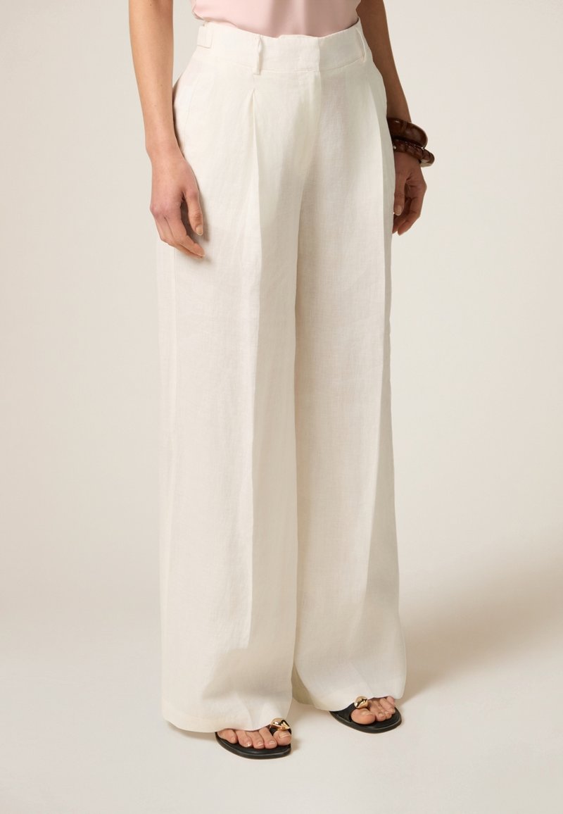 Witte linnen wijde broek met plooien, met een hoge taille en een lichte textuur, gecombineerd met zwarte sandalen met gouden accenten.