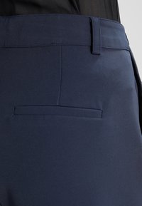 Detailní záběr na tmavě modré chinos kalhoty, které mají kapsu na zadní straně a poutko na opasek, nošené s zasunutým černým topem.