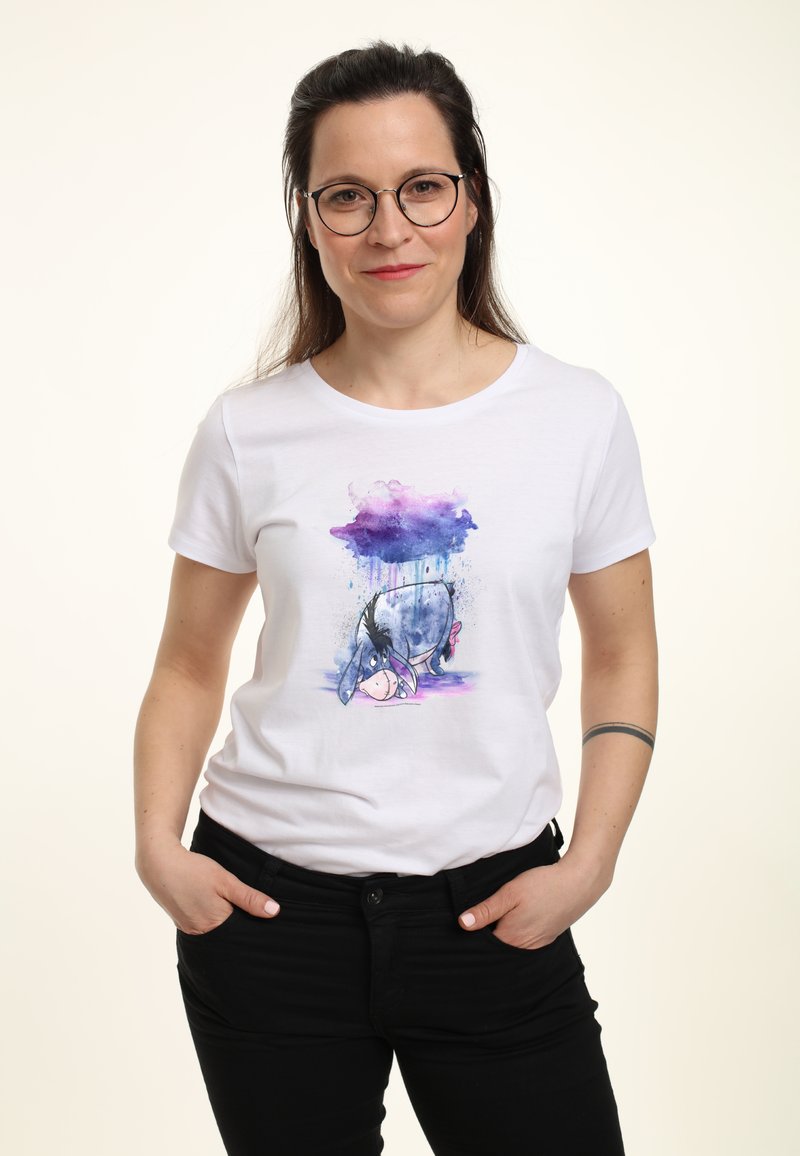 Camiseta blanca de algodón con un gráfico colorido de Eeyore y acentos en acuarela azul y púrpura. Modelo con pantalones negros, manos en los bolsillos.