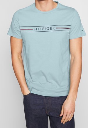 Mężczyzna w jasnoniebieskiej koszulce z krótkim rękawem z napisem "HILFIGER" i czerwono-biało-czarnymi paskami na piersi, w ciemnych dżinsach.