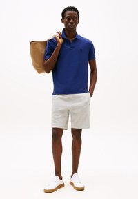 Junger Mann steht da und trägt ein blaues Poloshirt, hellgraue Shorts, weiße Sneakers und hält eine braune Tragetasche über einer Schulter.