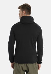 Black technical sweatshirt with a high collar, long sleeves, and flat seams, paired with olive green pants in a neutral setting.
