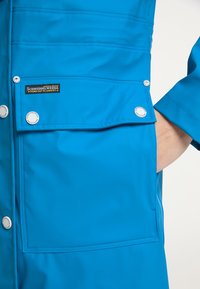 Voděodolná modrá bunda vyrobená z hladkého materiálu, s kapsou na hrudi se zaklapávacími knoflíky a značkovým štítkem na levé straně.