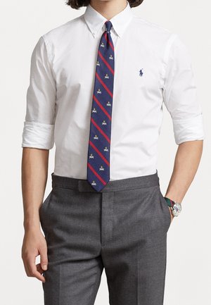 Biała koszula zapinana na guziki z podwiniętymi rękawami, granatowy krawat z czerwonymi skośnymi paskami i żółtymi akcentami, zestawiony z szarymi spodniami.