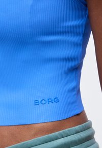 Κοντινό πλάνο ενός μπλε ριμπ crop top με το λογότυπο «BORG» ανάγλυφο κοντά στο τελείωμα, που φοριέται από άτομο με σκουρόχρωμο δέρμα και πράσινο παντελόνι.